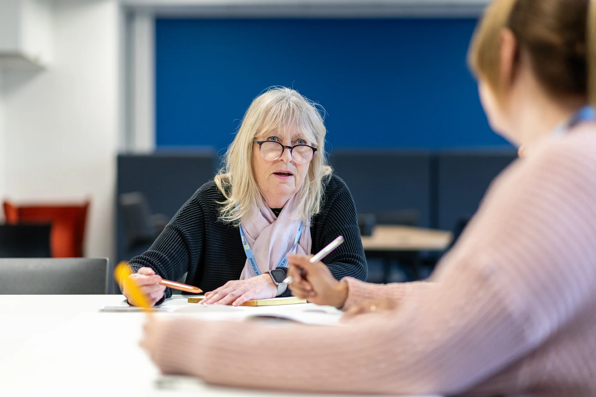 Middle aged lady sat at a desk writong notes and talking to another lady